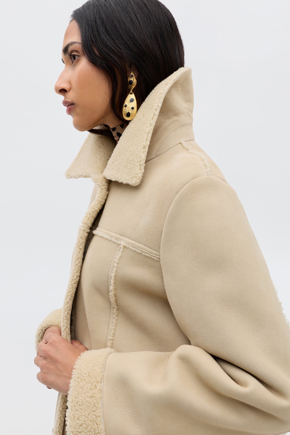 A woman in profile with dark hair and gold statement earrings wears the Ellis - Cream faux-shearling coat, featuring a wide collar and cuffs, holding the lapel against a plain light background.
