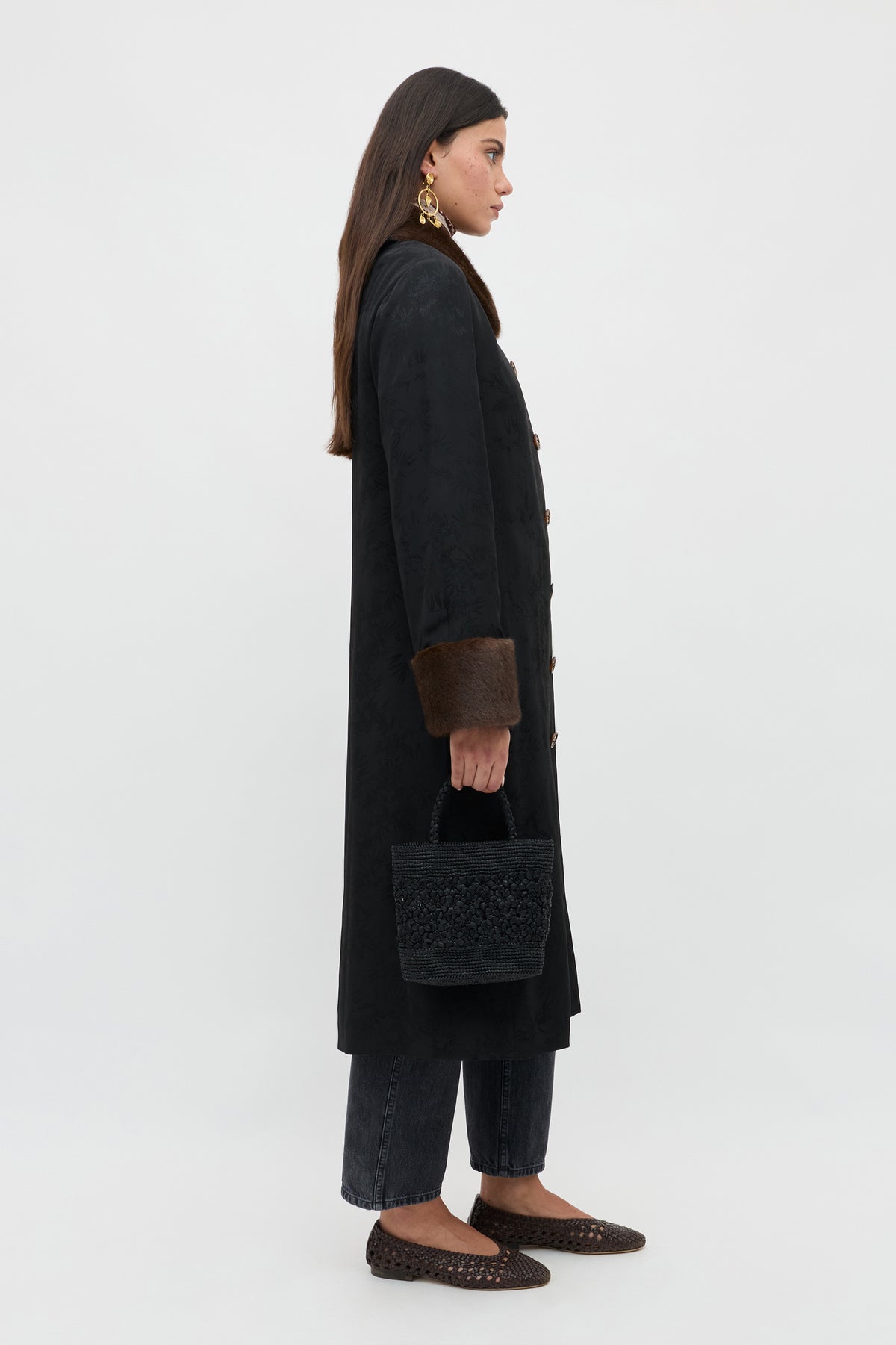 A woman stands in profile against a plain background, wearing a long black coat with brown fur cuffs, dark pants, brown shoes, gold earrings, and holding the Jonah - Black textured handbag.