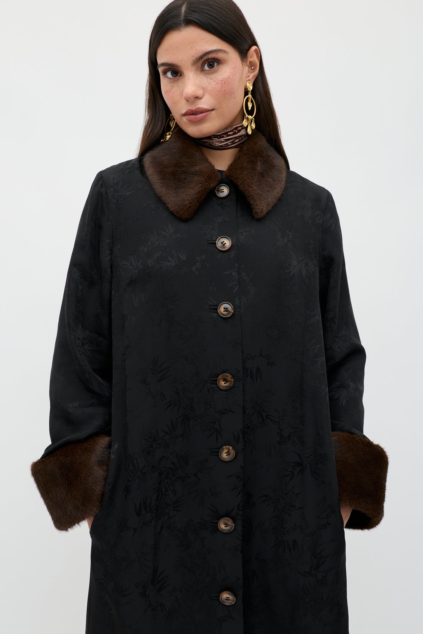A woman wearing the Jonah - Black coat with brown fur collar and cuffs, gold earrings, and a patterned neck scarf stands against a plain light background.