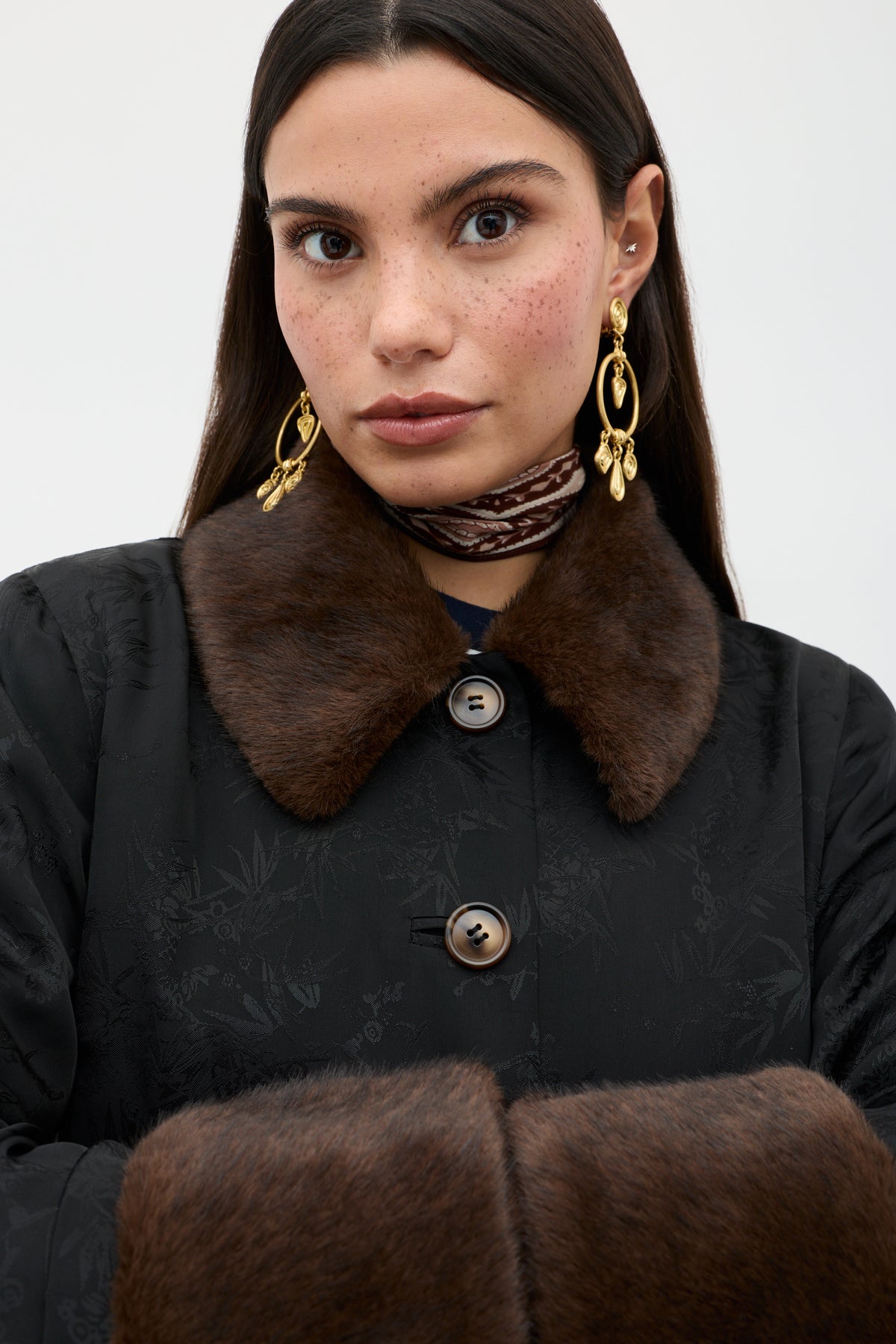 A woman with long dark hair and freckles wears the Jonah - Black coat featuring a brown fur collar and cuffs, gold earrings, and a patterned scarf, standing against a plain light background.
