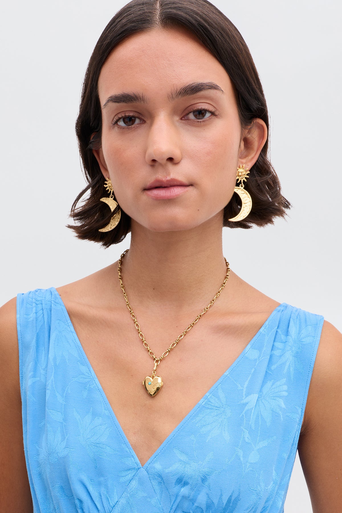A woman with short brown hair wears the Veyra - Gold crescent moon earrings and a cosmic-inspired locket. She is dressed in a light blue sleeveless floral dress, standing against a plain background.