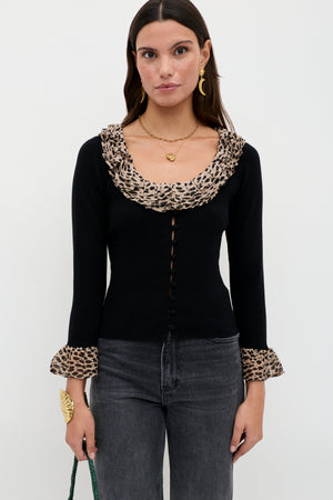 A woman with long brown hair and gold jewelry models the Laurel - Antelope Spot Black cardigan, featuring leopard-print collar and cuffs, paired with dark jeans and a green bag against a plain white background.