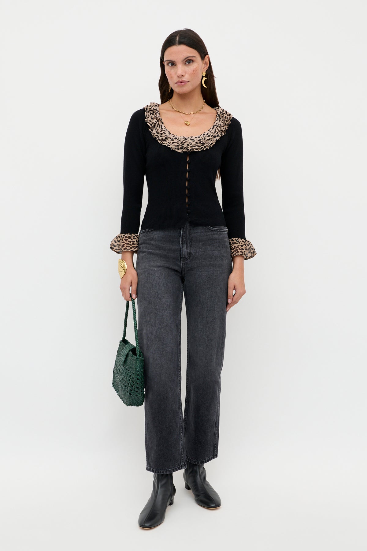 A woman stands against a white background wearing the Laurel - Antelope Spot Black cardigan with leopard-print trims, gray jeans, black ankle boots, gold jewelry, and holding a green woven handbag.