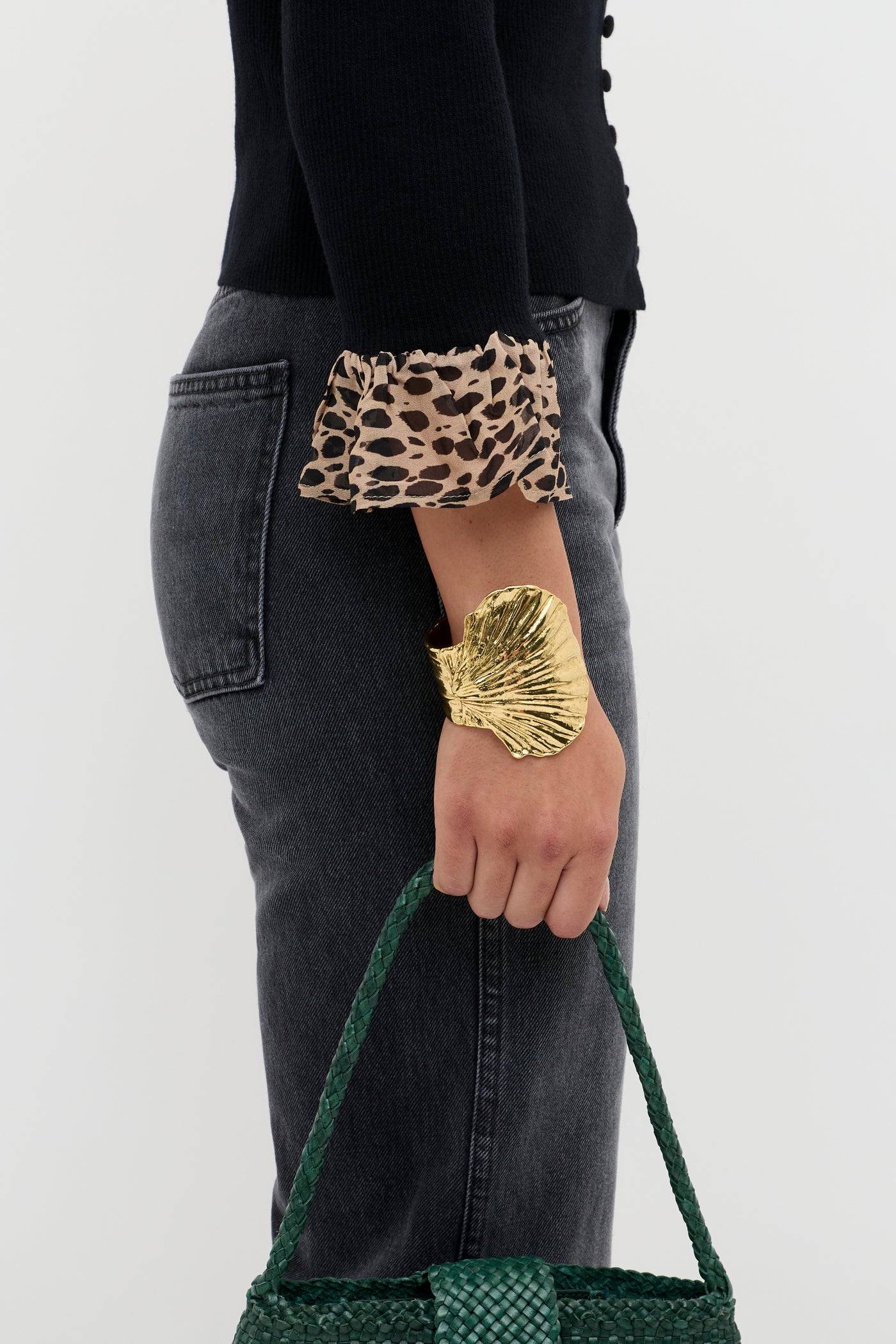 A person wearing dark jeans and a black cardigan with leopard-print trims holds the Laurel - Antelope Spot Black green woven handbag and wears a large gold leaf-shaped bracelet.