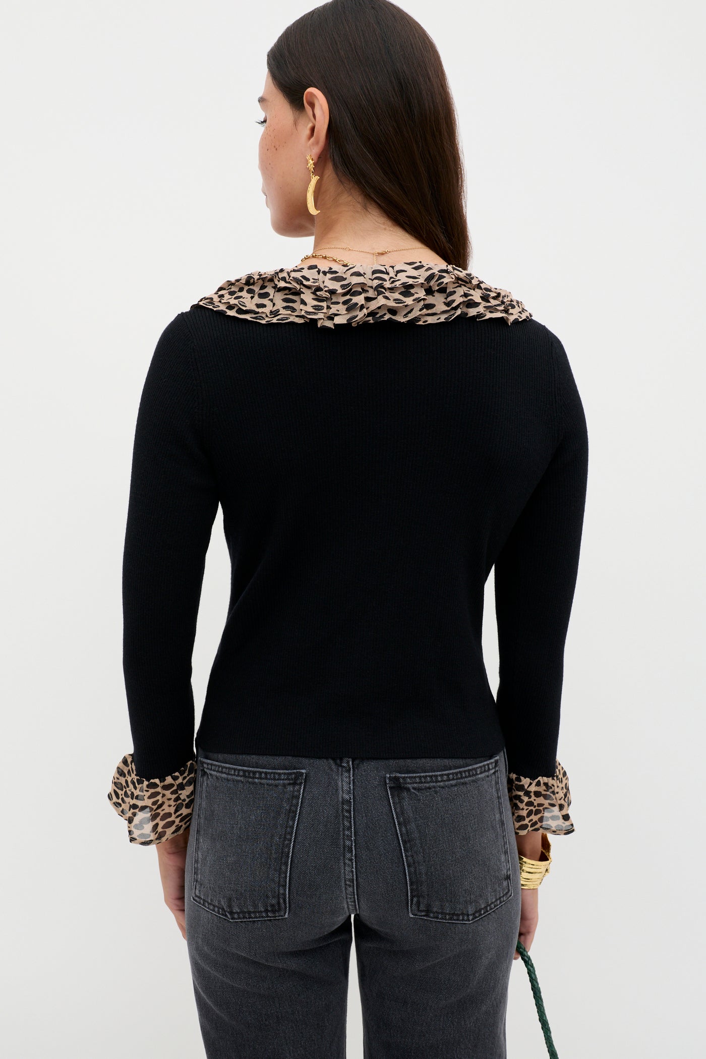 A woman with long brown hair stands facing away from the camera against a plain light background, wearing the Laurel - Antelope Spot Black cardigan with leopard-print collar and cuffs, paired with high-waisted dark gray jeans.