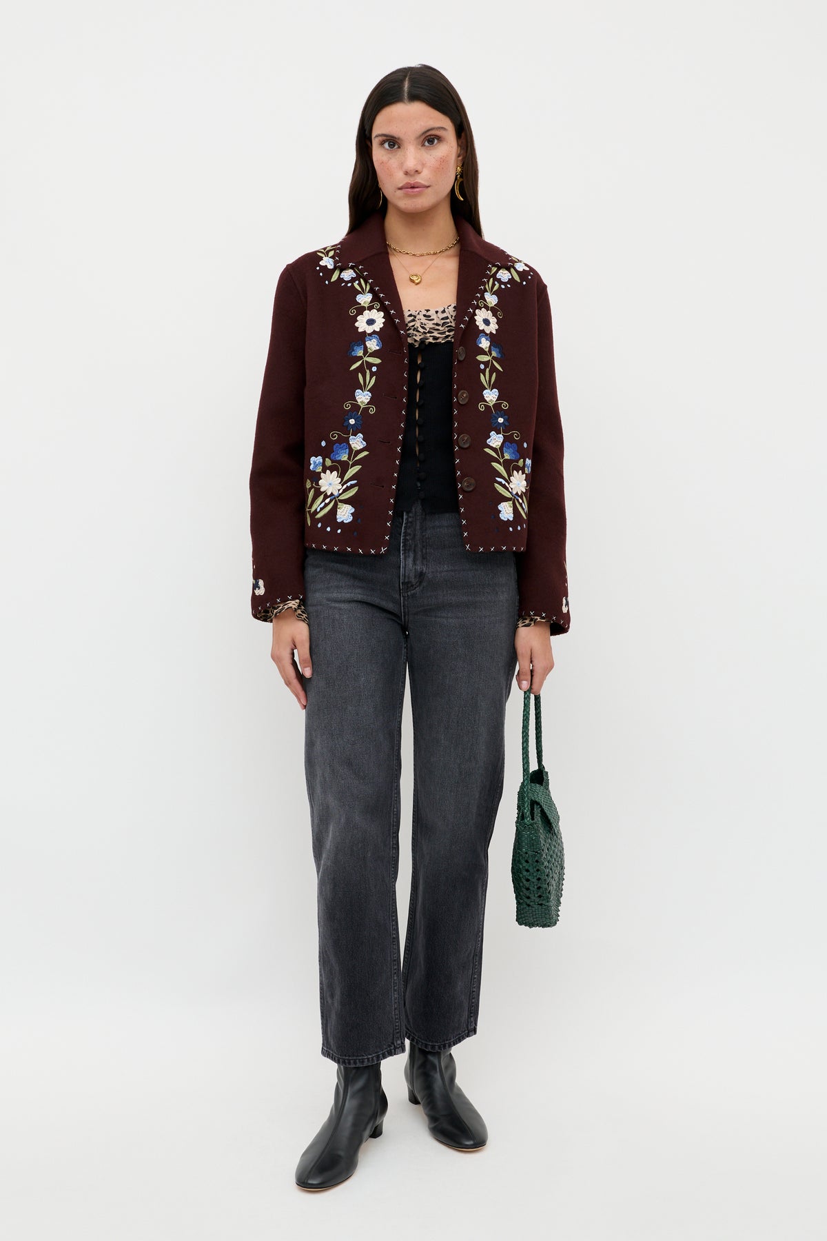 A woman poses against a white background wearing the Laurel - Antelope Spot Black jacket, which features floral embroidery and subtle leopard-print trims, styled with a black top, gray jeans, black ankle boots, and a green woven handbag.