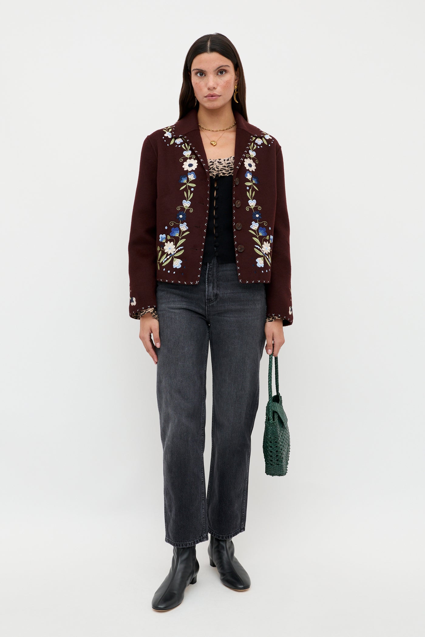 A woman poses against a white background wearing the Laurel - Antelope Spot Black jacket, which features floral embroidery and subtle leopard-print trims, styled with a black top, gray jeans, black ankle boots, and a green woven handbag.