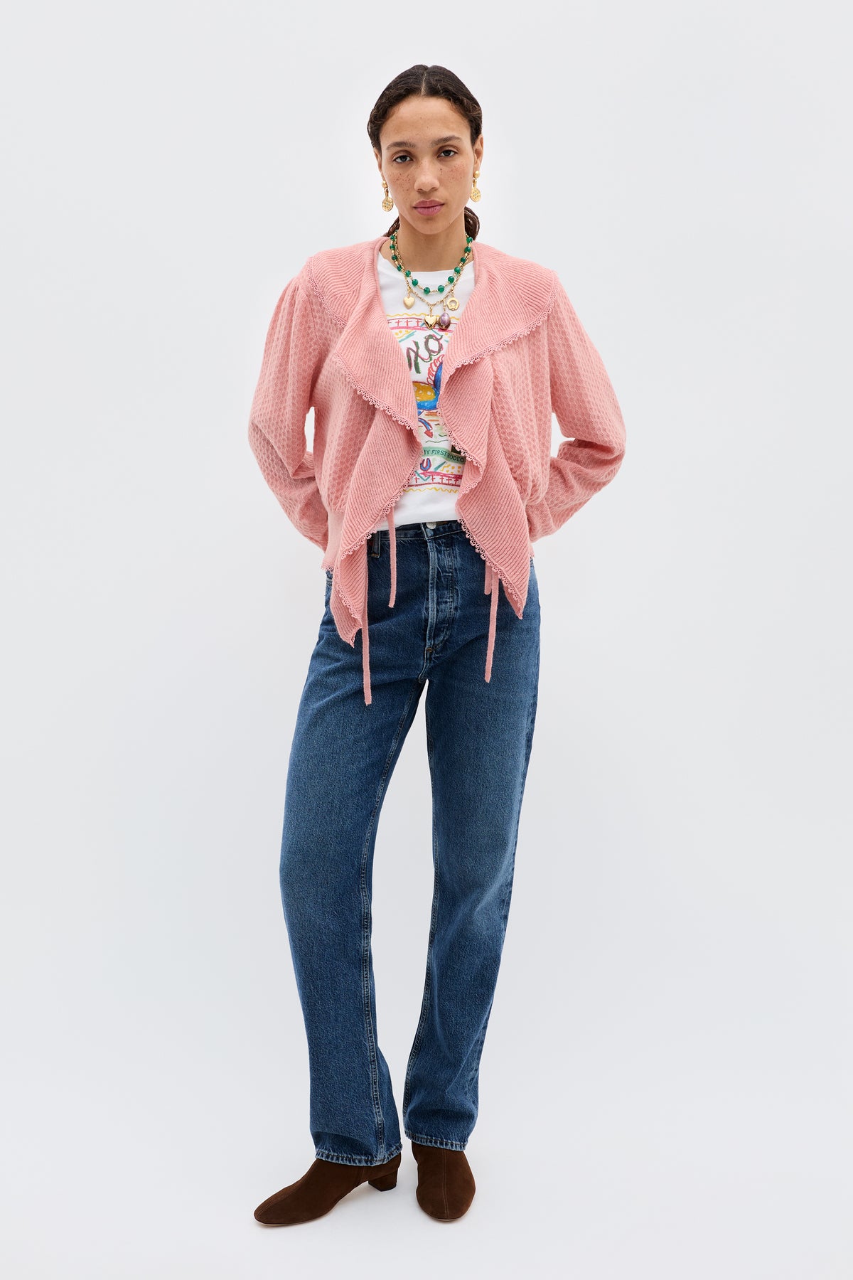 A person wearing the Kianna - Blush alpaca wool blend cardigan over a graphic tee, blue jeans, and brown shoes stands against a plain white background with hands in their pockets.