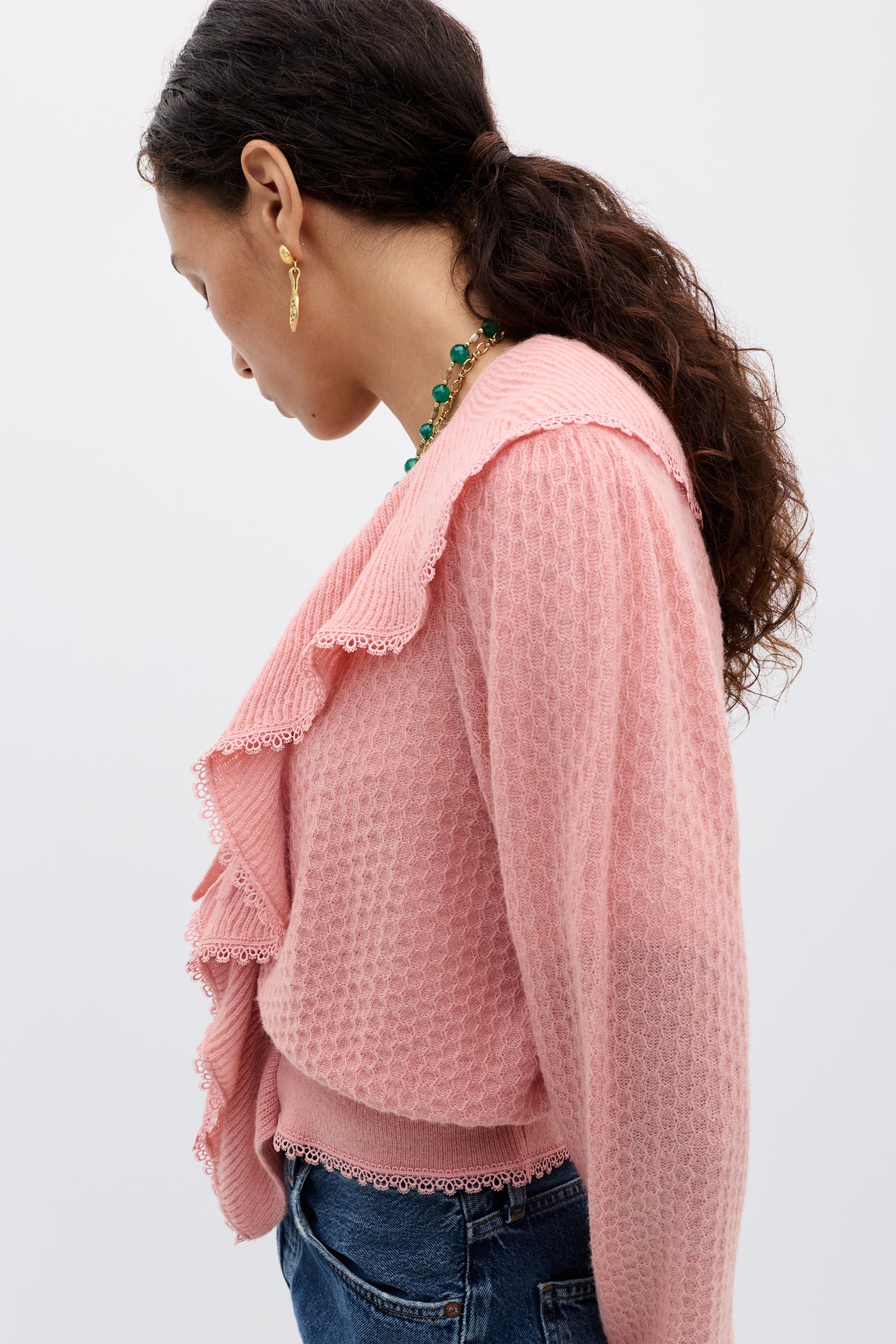 A woman with wavy brown hair in a ponytail wears the Kianna - Blush knit cardigan, blue jeans, gold earrings, and a green beaded necklace, standing in side profile against a plain light background.