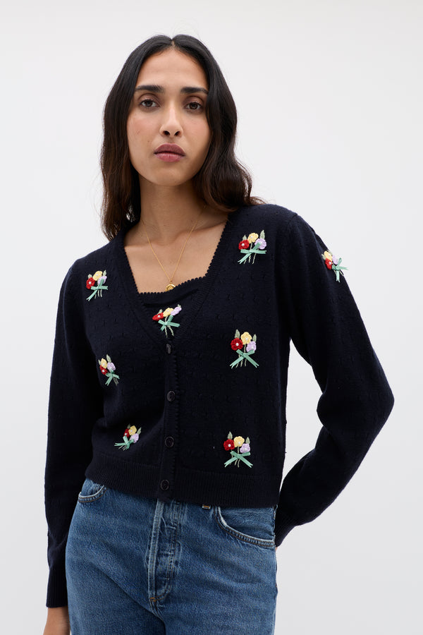 A woman with long dark hair wears the Merinda - Floret Embroidery Deep Navy cardigan, featuring soft merino wool and colorful floral embroidery, paired with blue jeans against a plain light background.