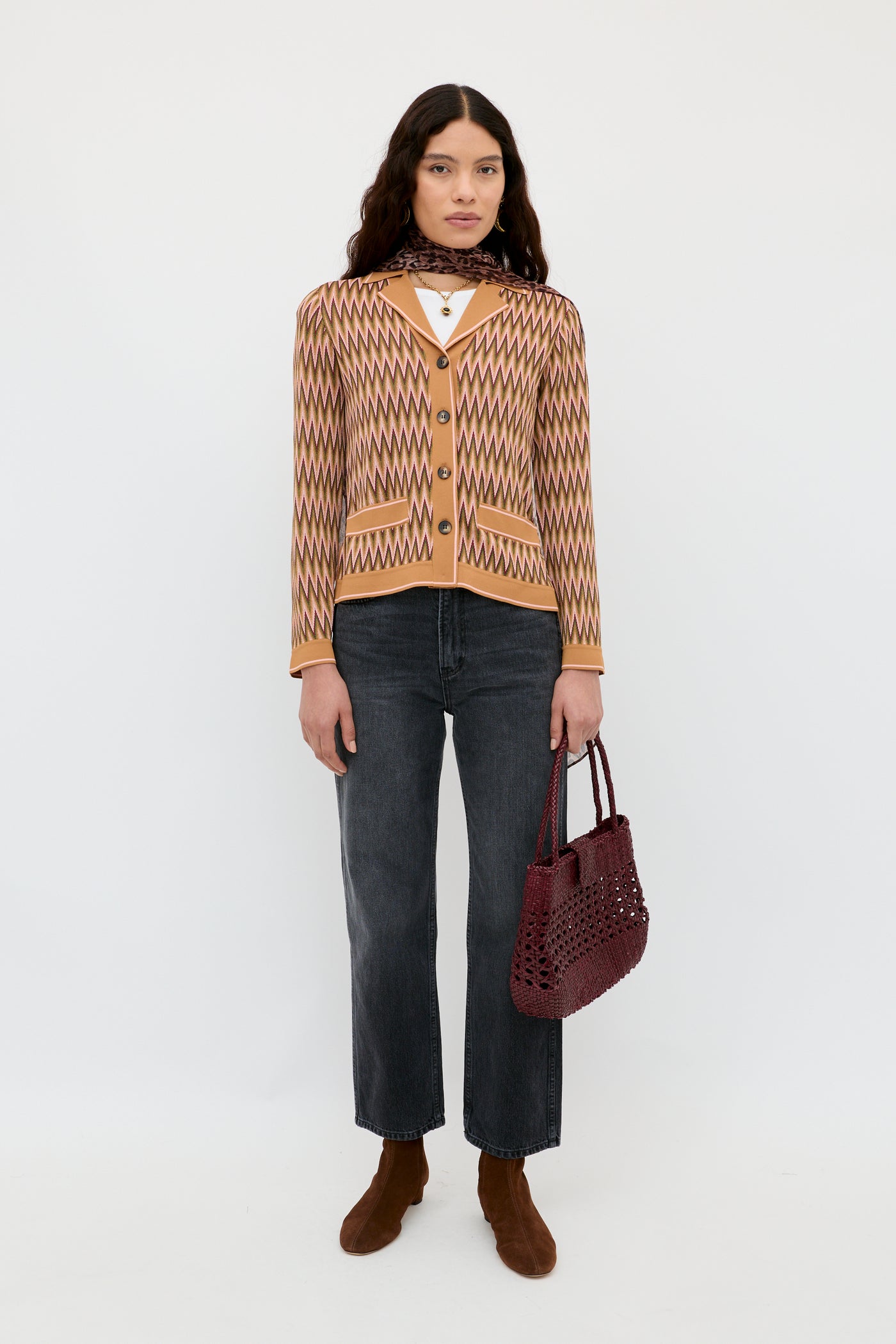 A woman with long dark hair and a neutral expression stands against a white background wearing the Quinley Maeve Jacquard Multi cardigan, paired with dark jeans, brown ankle boots, and holding a burgundy woven handbag.