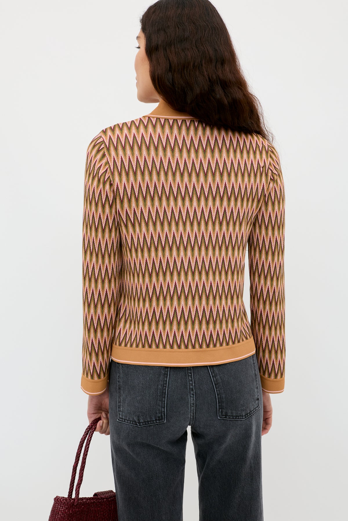 A person with long, wavy dark hair wears the Quinley - Maeve Jacquard Multi sweater in beige, brown, and pink zigzag patterns, black jeans, and holds a maroon woven bag against a plain light background.