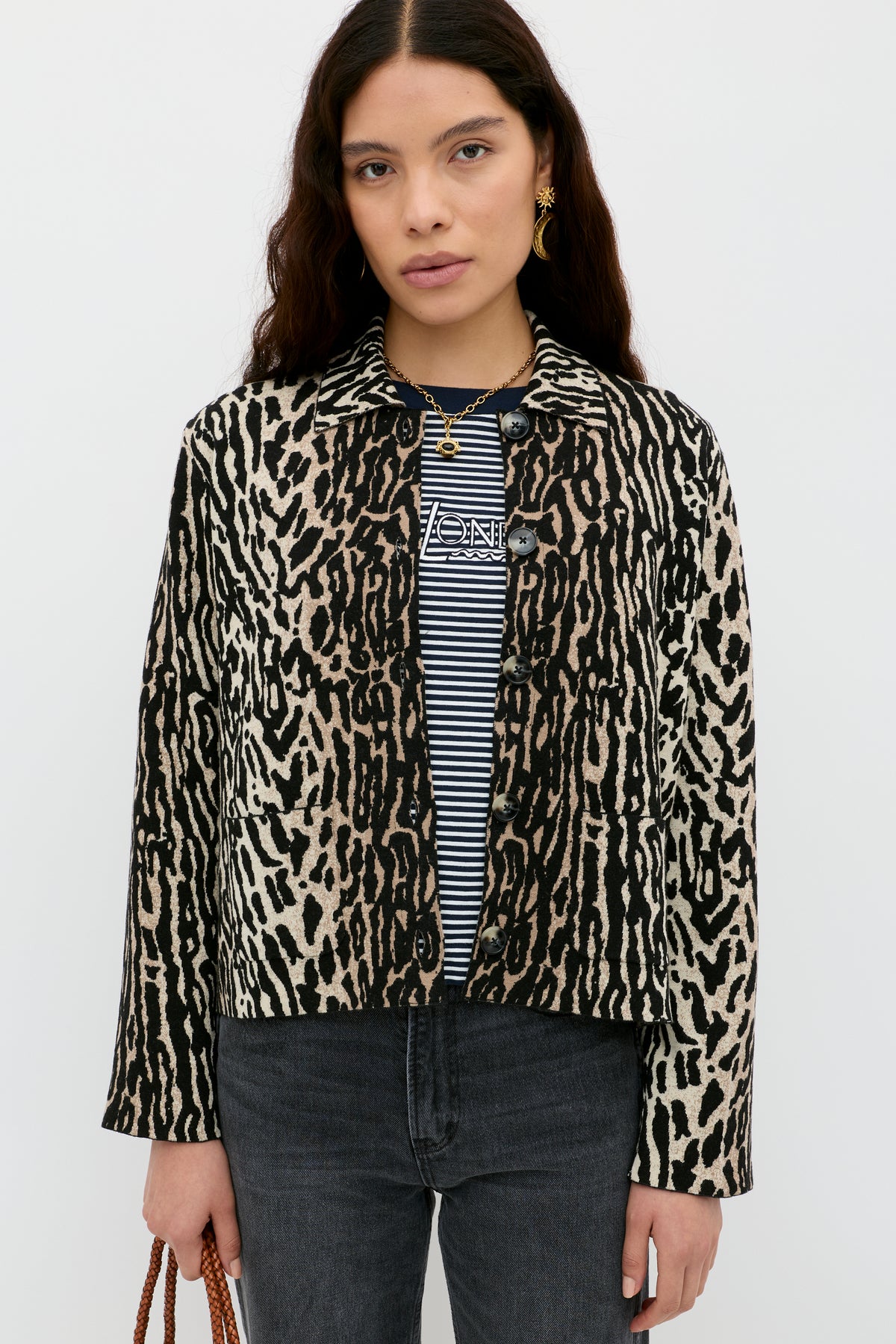 A woman with long dark hair stands against a white background, wearing the Trista - Bohemia Leopard Brown jacket over a black-and-white striped shirt and black jeans. She holds a brown bag in her left hand.
