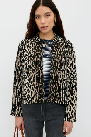 A woman with long dark hair stands against a white background, wearing the Trista - Bohemia Leopard Brown jacket over a black-and-white striped shirt and black jeans. She holds a brown bag in her left hand.