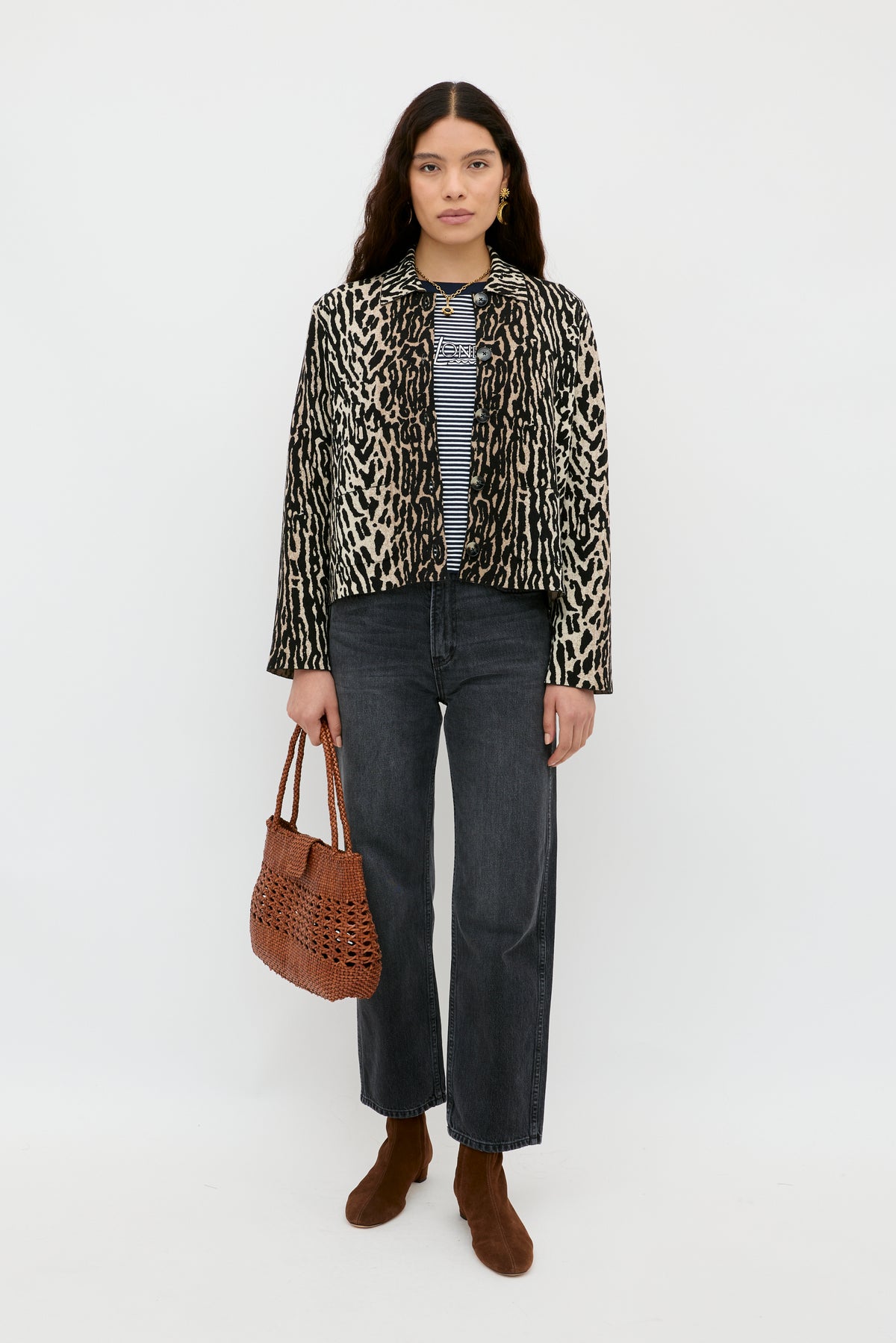 A woman with long dark hair models the Trista – Bohemia Leopard Brown jacket over a striped shirt, dark jeans, brown ankle boots, and a brown woven handbag against a plain white background.
