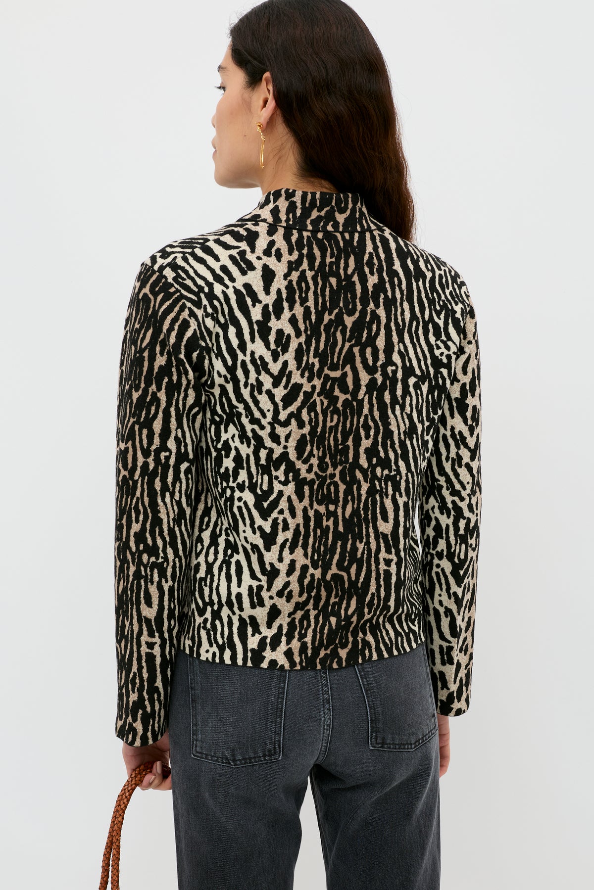 A woman with long dark hair, wearing a black and beige animal print jacket and black jeans, stands facing away from the camera against a white background. She holds the Trista - Bohemia Leopard Brown by its brown strap in her left hand.