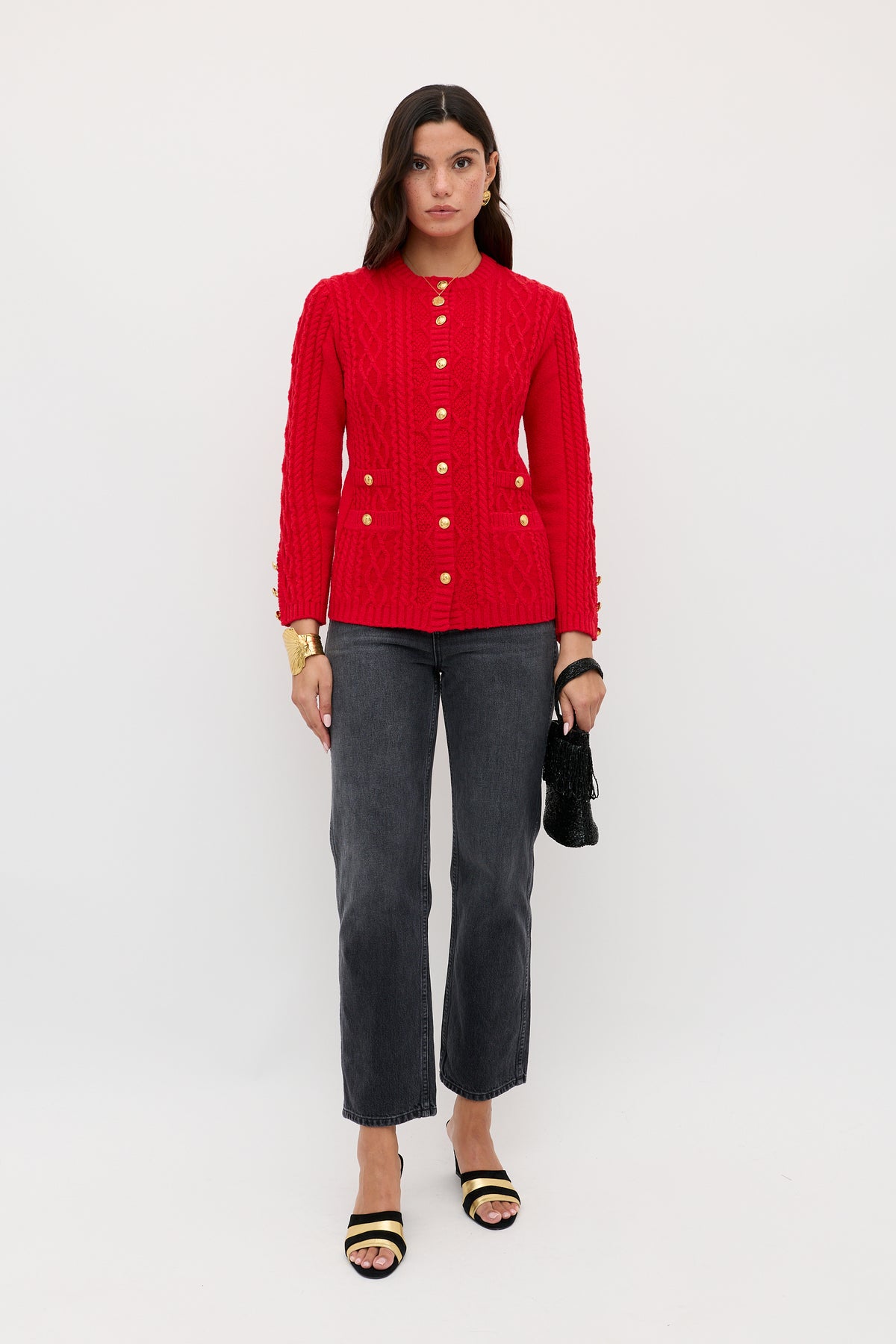 A woman stands against a plain white background wearing the Maurya - Red textured button-up jacket, dark gray straight-leg jeans, black and gold sandals, a chunky gold bracelet, and holding a small black clutch.