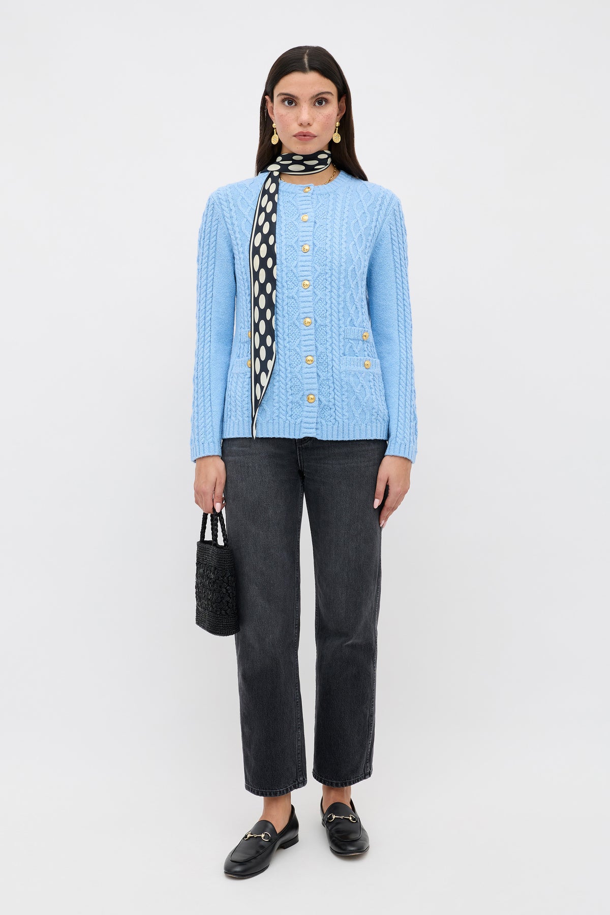 A woman faces forward in a Maurya - Dusty Blue cable knit cardigan, black and white polka dot scarf, dark gray jeans, black loafers, and a small black handbag against a plain white background.