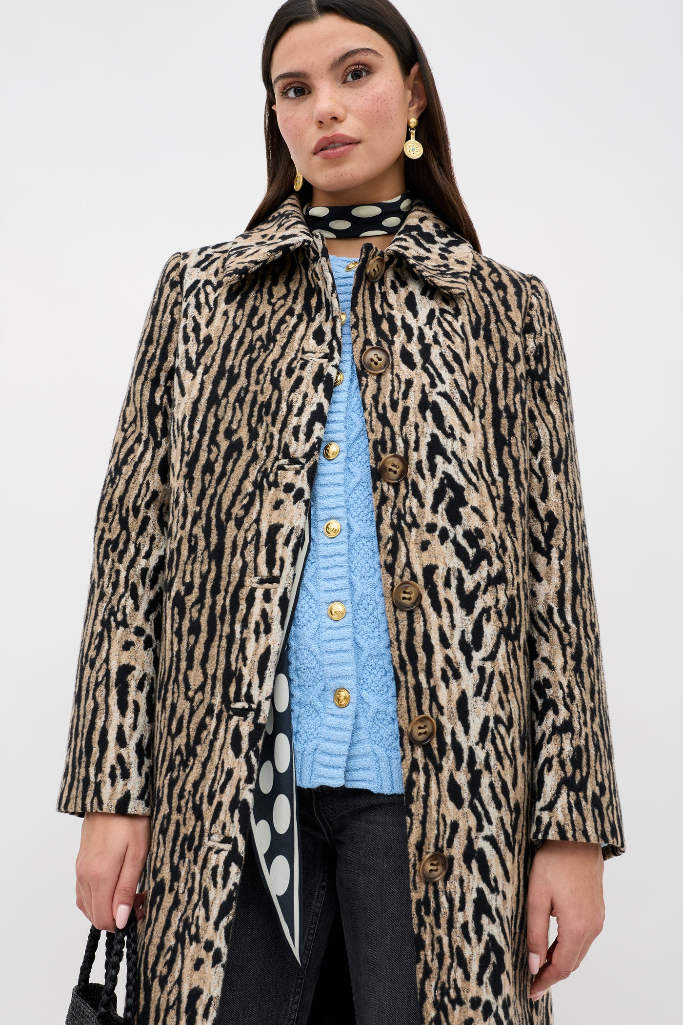 A woman wears a leopard print coat over the Maurya - Dusty Blue sweater, paired with black jeans and a black scarf with large white polka dots. She has straight brown hair, gold earrings, and carries a textured black bag.