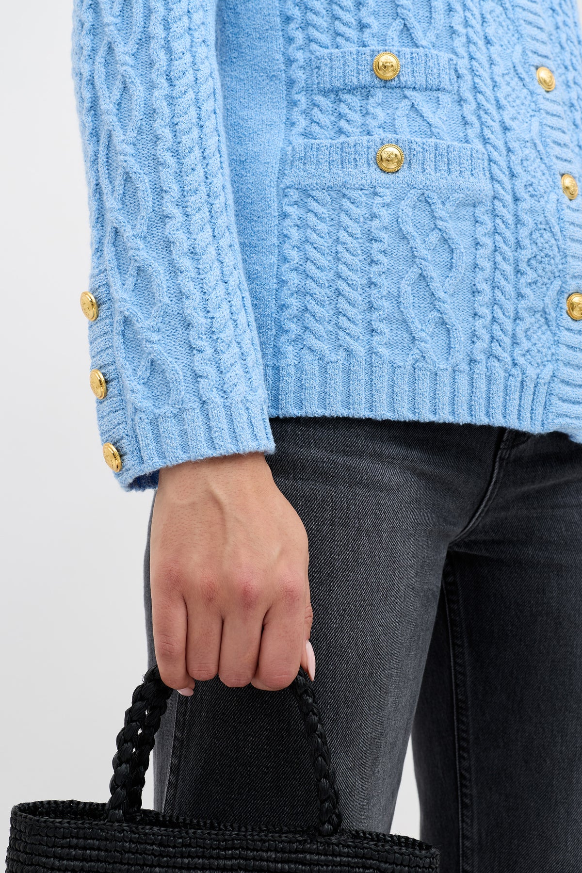 Wearing the Maurya - Dusty Blue cable knit sweater, paired with dark gray jeans, a person holds a black woven handbag. The image highlights the lower half of the sweater, hand, and bag.
