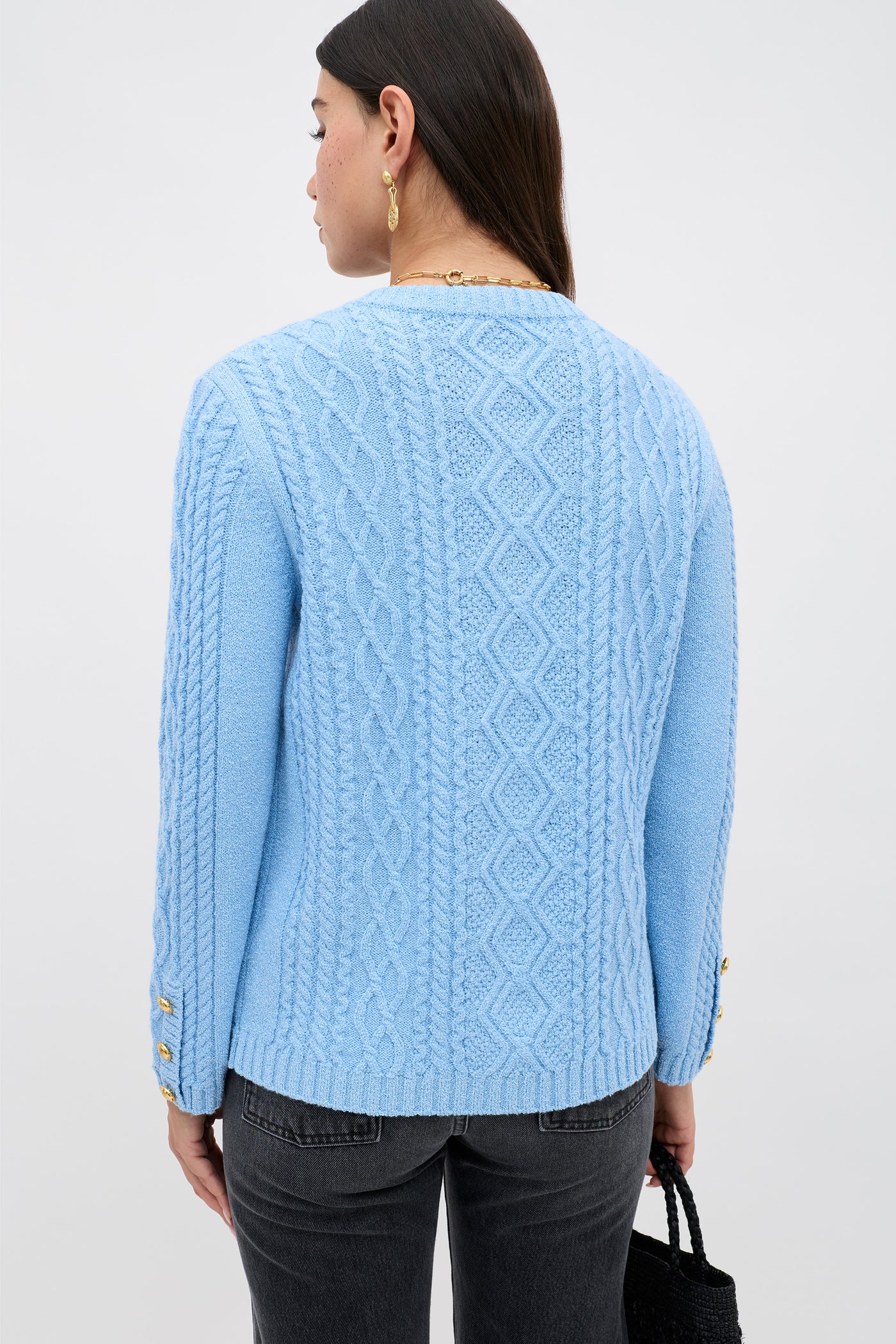 A person with long dark hair, gold earrings, and dark jeans is shown from the back against a plain light background, wearing the Maurya - Dusty Blue cable-knit sweater with diamond and braid patterns.