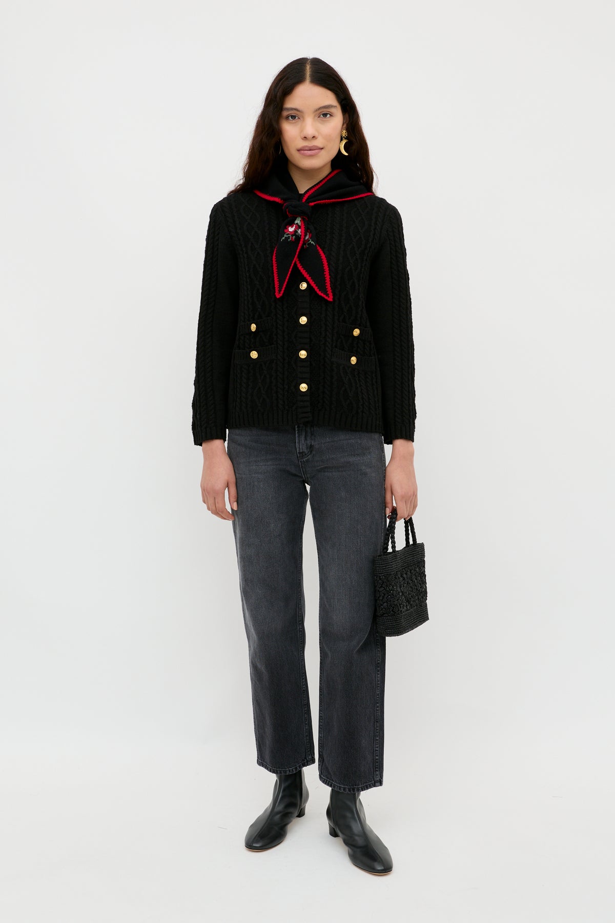 A woman stands against a white background wearing the Maurya - Black textured jacket with gold buttons, a red-trimmed scarf, dark gray jeans, black ankle boots, and holding a small black bag.