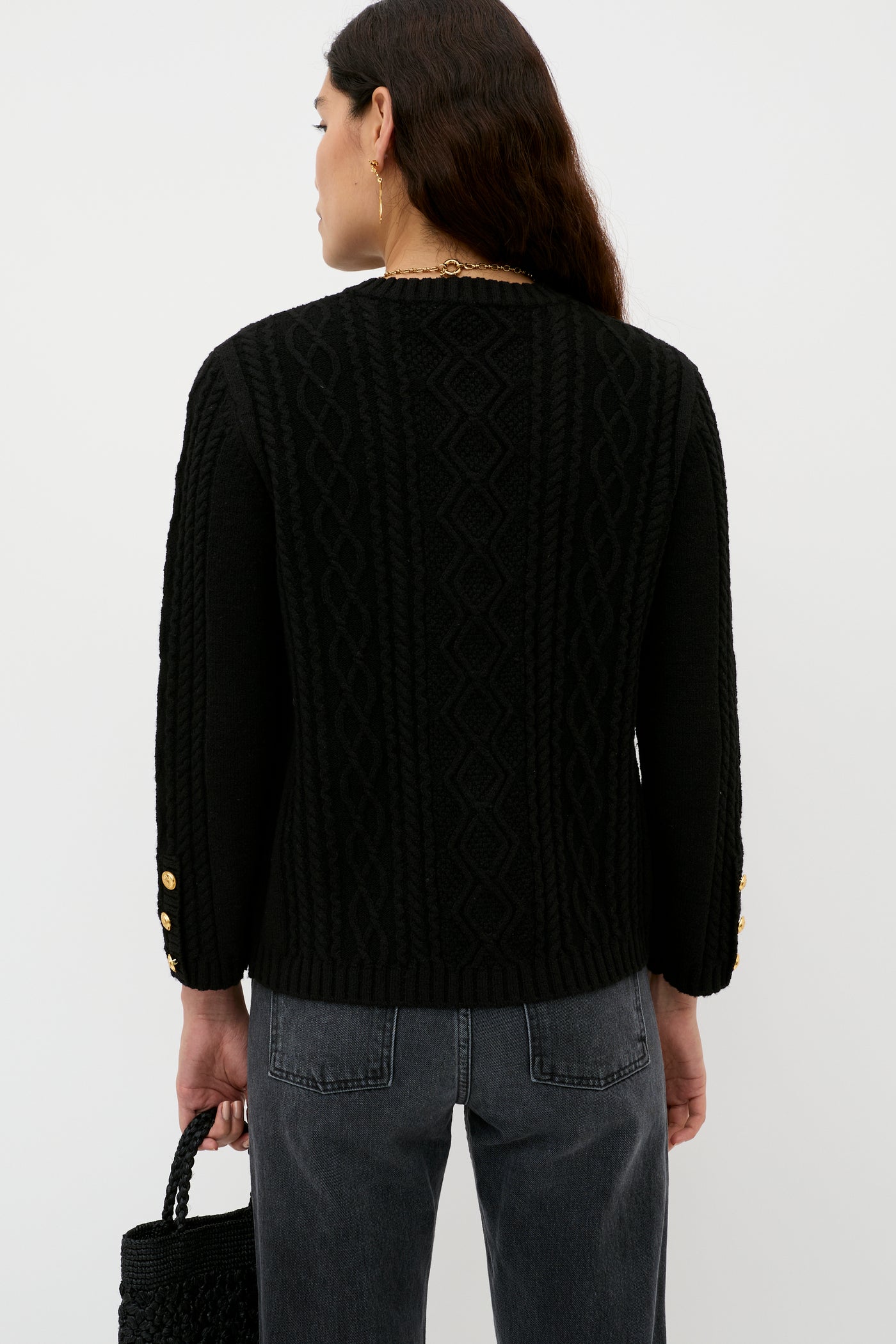 A person with long dark hair, wearing a textured black sweater with gold sleeve buttons and dark jeans, stands facing away holding the Maurya - Black woven bag against a plain white background.
