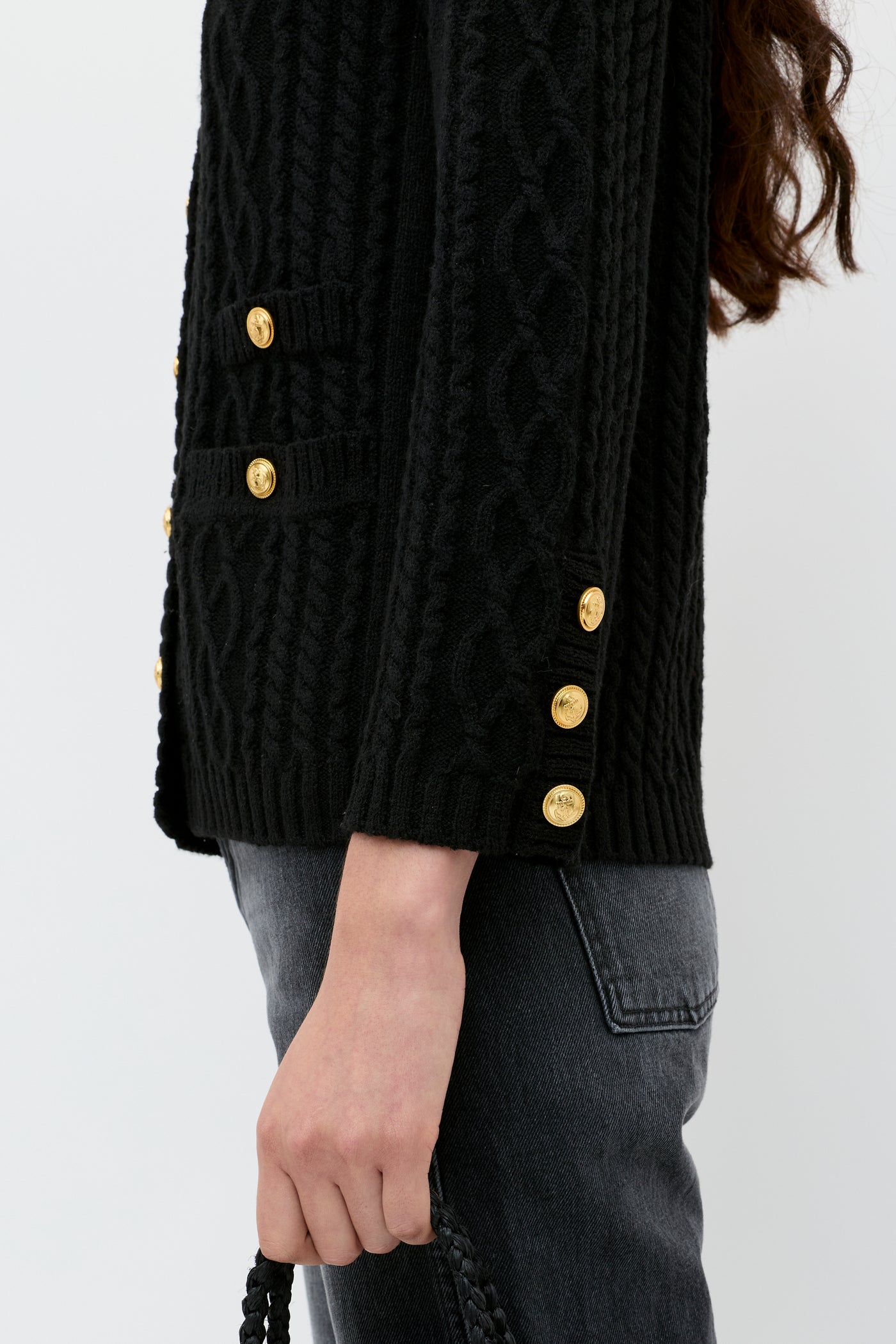 A person is wearing dark jeans, a black cable knit sweater with gold buttons, and holding the Maurya - Black braided strap handbag. The photo is cropped to show only their torso and part of one arm.