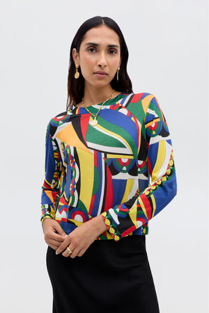 A woman with long dark hair, wearing gold jewelry and the Phaedra - Soho Geo Multi crop top with a black skirt, stands against a plain light background.