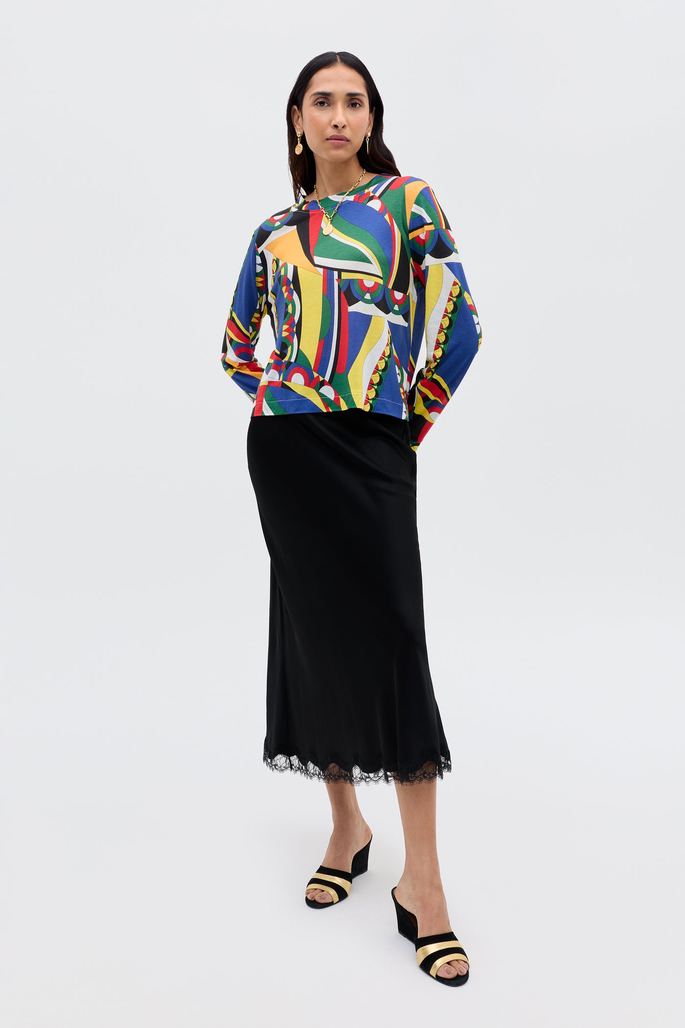 A person stands facing forward, wearing the Phaedra - Soho Geo Multi long-sleeve top in soft Tencel Jersey, paired with a black midi skirt with lace hem, black open-toe heels, and gold earrings against a plain light background.