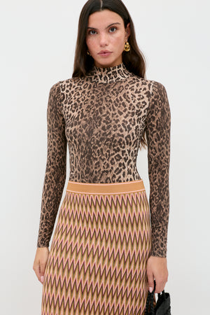 Woman with long brown hair wears the Polo - Leopard Brown bodysuit paired with a high-waisted, beige skirt featuring a wavy zigzag pattern, standing against a plain white background.