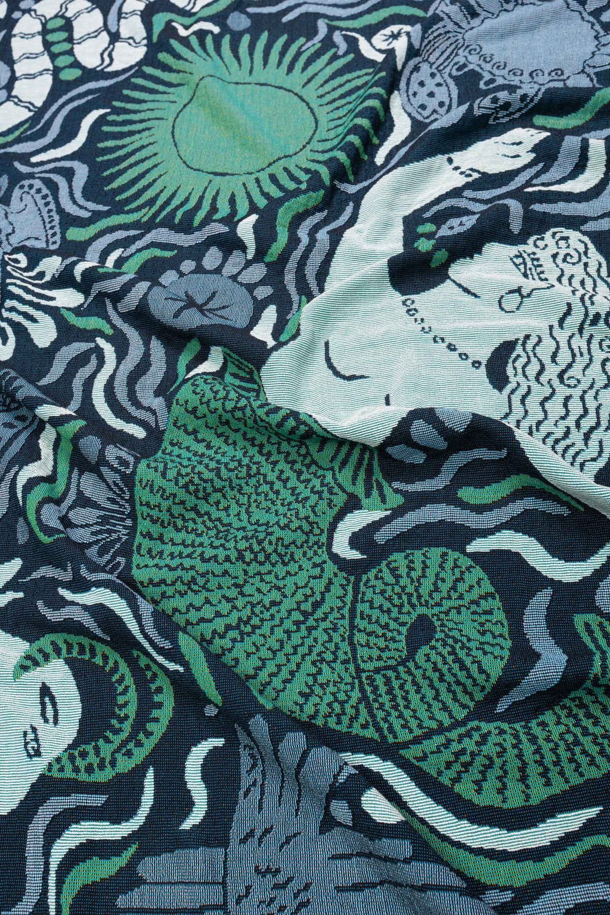 A close-up of the Mythos Throw - Navy from the Mythos Collection highlights its woven texture and intricate design, featuring abstract sea creatures, a mermaid, shells, and flowing lines in shades of green, blue, and teal. Inspired by Greek mythology, the detailed craftsmanship is beautifully visible.