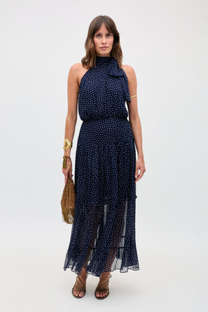 A woman stands against a plain background, wearing the Abi - Mini Navy Polka Dot dress with white polka dots. She accessorizes with a gold fringe handbag, gold jewelry, and brown strappy sandals.