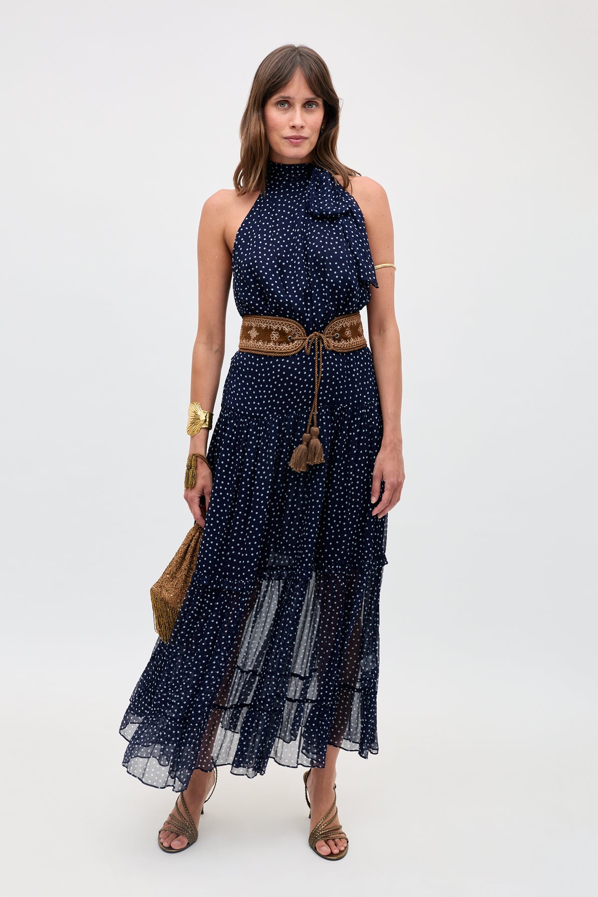 A woman in the Abi - Mini Navy Polka Dot dress, styled sleeveless with a high neckline, accessorized with a brown belt, gold jewelry, tan sandals, and a woven bag, poses against a plain light background.