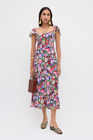 A woman wearing the Adele - Ikat Bloom colorful floral midi dress, statement earrings, and sandals, holding a woven handbag, stands against a plain white background.