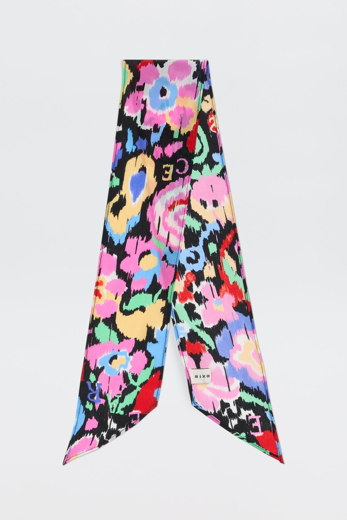 The Alexa - Ikat Bloom Charcoal is a silk scarf with abstract floral patterns in pink, yellow, blue, green, and red on a black background. It features pointed ends, a small brand label, and is displayed against a white backdrop.