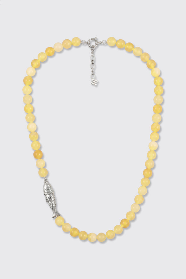 The Amoura - Lemon necklace is made of round yellow beads and features a silver fish-shaped charm on one side, along with a silver clasp and an adjustable chain.