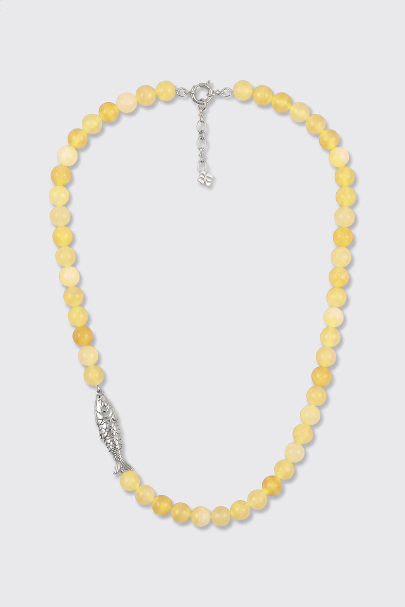 The Amoura - Lemon necklace is made of round yellow beads and features a silver fish-shaped charm on one side, along with a silver clasp and an adjustable chain.
