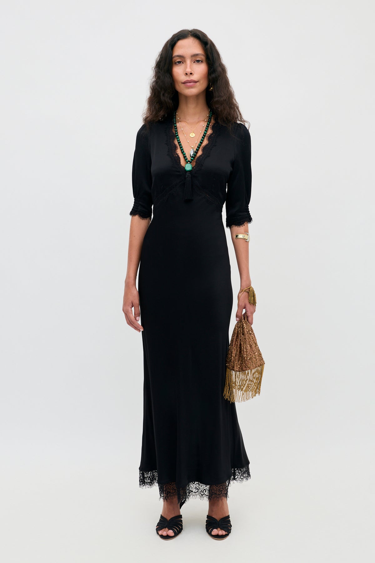 A woman wearing the Annina - Black midi dress with lace trims, cut on the bias for a flattering silhouette, paired with black sandals, layered necklaces, a gold bracelet, and holding a gold fringe handbag, stands against a plain white background.