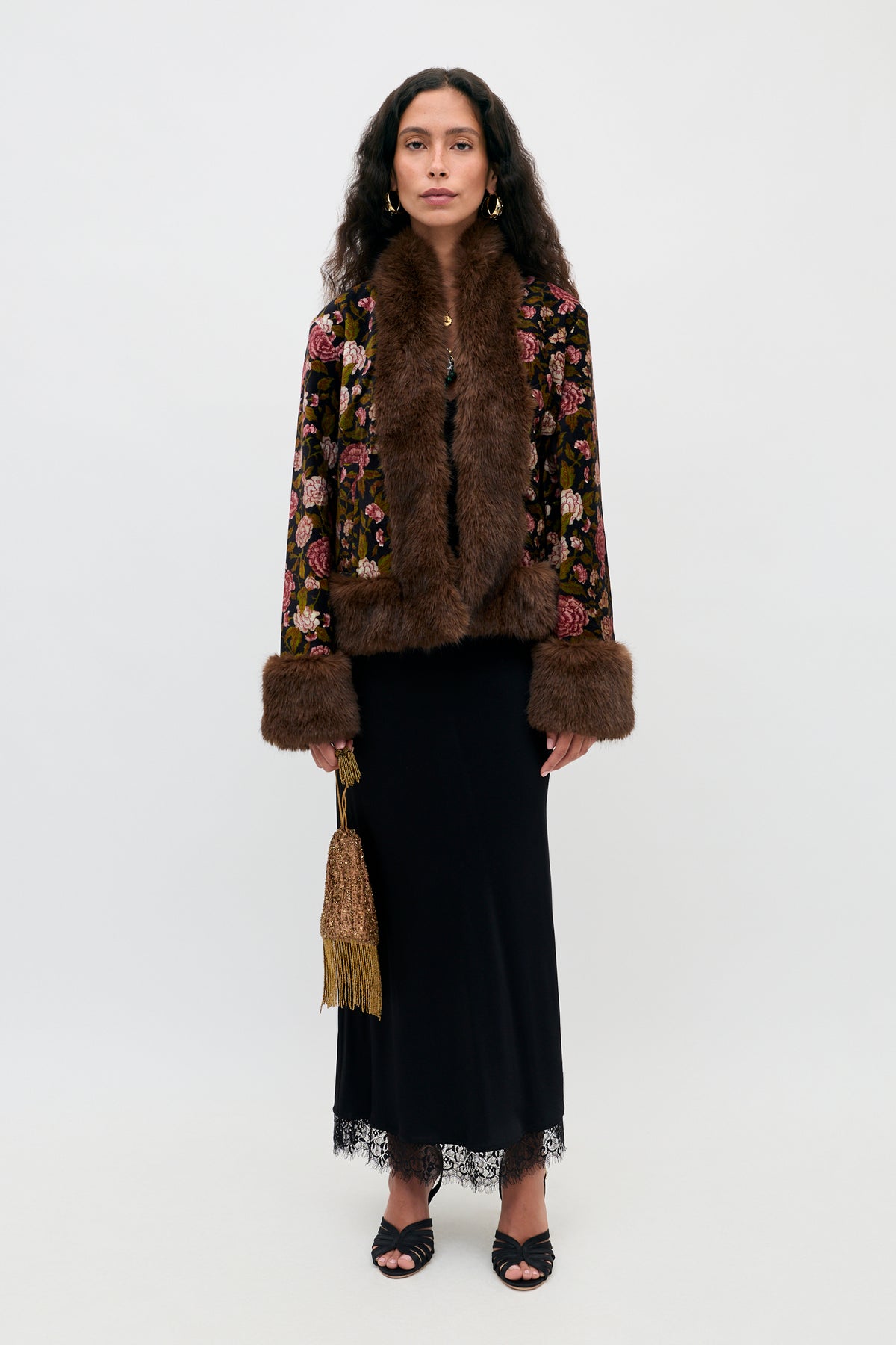 A woman stands against a white background wearing the Annina - Black midi dress, which features delicate lace trims at the hem and a flattering bias cut. She pairs it with a floral jacket with brown fur trim, black sandals, and holds a gold fringed purse.