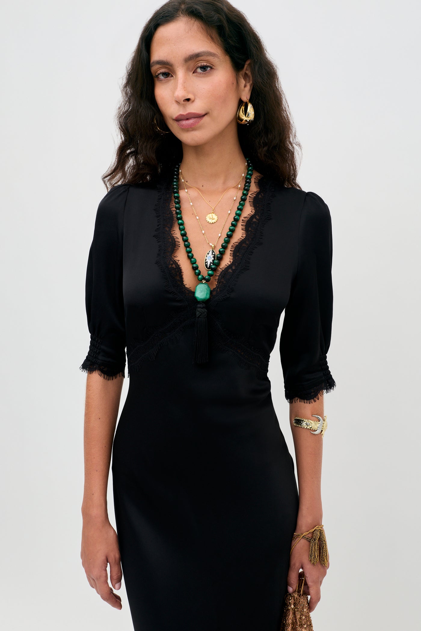 A woman with long curly hair wears the Annina - Black midi dress, featuring a deep V neckline, bias cut silhouette, and delicate lace trims. She accessorizes with gold earrings, layered necklaces, and gold bracelets while standing against a light background.