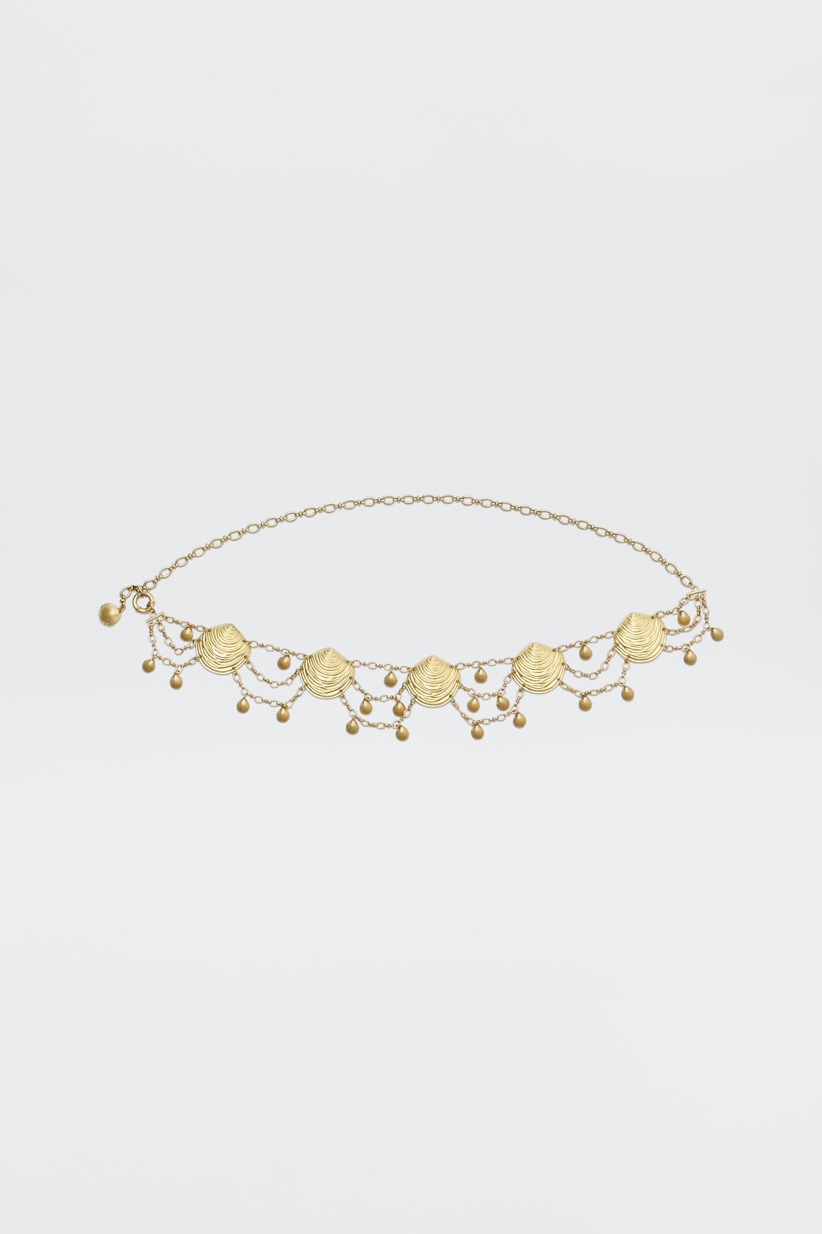 The Asia - Gold necklace is a delicate gold piece with small scallop shell charms and dangling beads symmetrically arranged on a fine chain, shown against a plain white background.