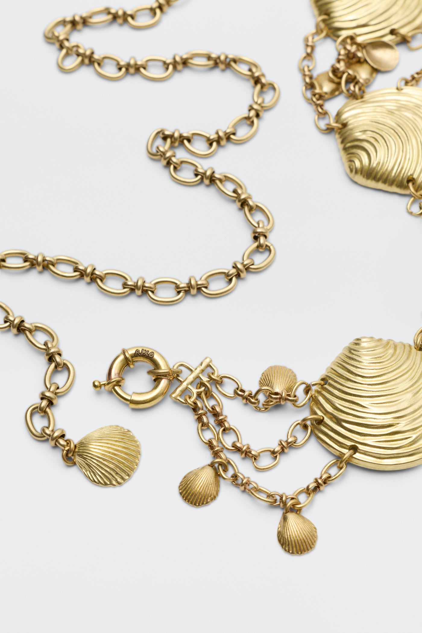 Asia - Gold necklace, featuring textured, shell-shaped pendants and decorative chain links with small dangling shells and a round clasp, is displayed laid flat on a white background.