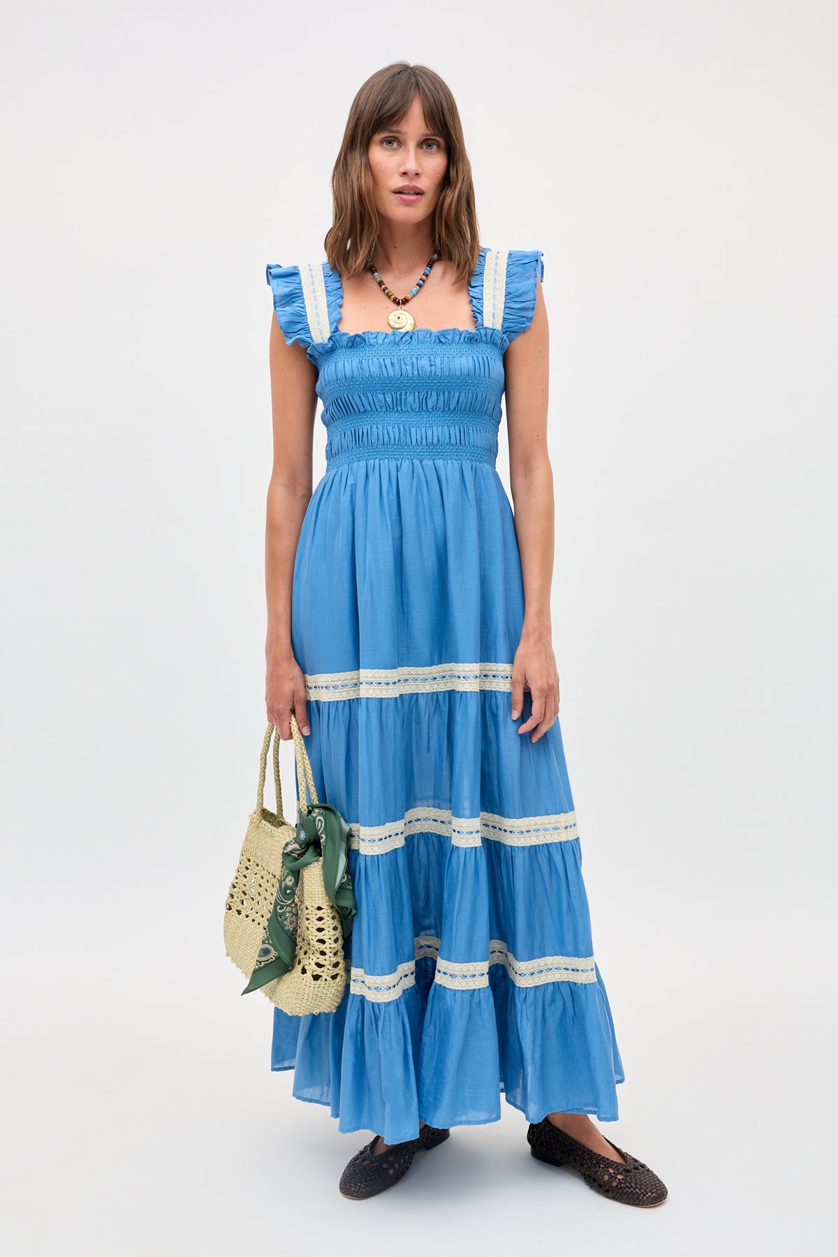 A woman wearing the Aubrie - Blue sleeveless maxi dress with ruffle straps and delicate lace trim stands holding a woven bag adorned with a green scarf. The elegant shirred panels of the dress enhance its flowing silhouette. She has straight brown hair and accessorizes with brown flats and a necklace.