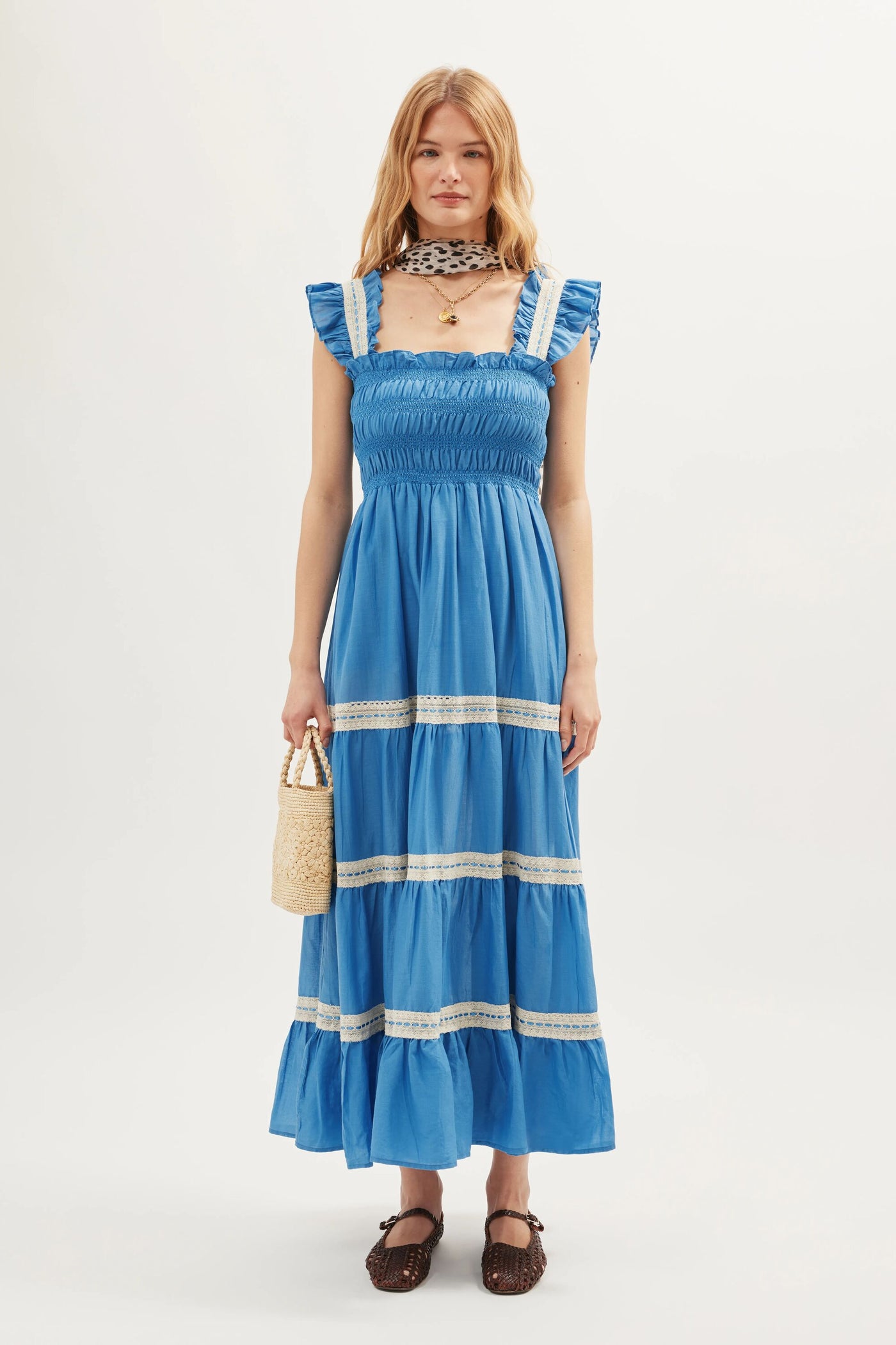 A woman with simply styled blonde hair stands against a white background, wearing the Aubrie - Blue ruffled maxi dress with delicate shirred panels and lace trim. She pairs it with brown shoes, a patterned neck scarf, and carries a small woven handbag.