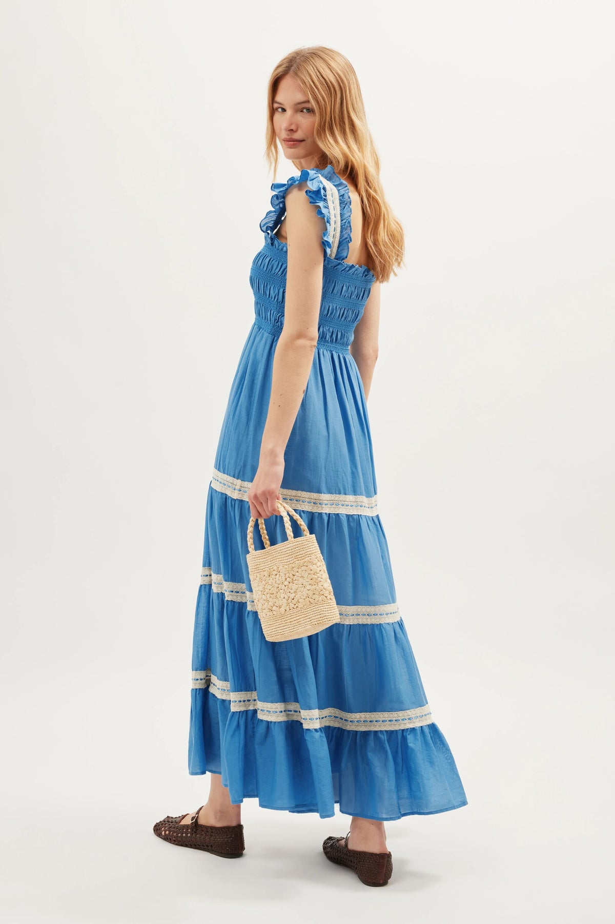 Wearing the Aubrie - Blue, an ankle-length dress featuring lace details and delicate shirred panels, the woman holds a small woven handbag. Her long, wavy hair cascades down as she wears brown woven shoes. She stands against a plain background, turned slightly and looking over her shoulder.