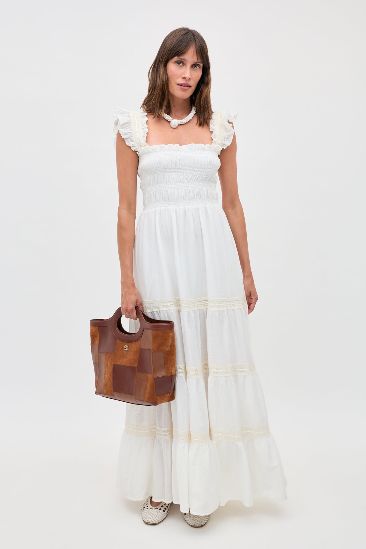 Woman in a long, 70s-inspired ivory nightdress called “Aubrie” with ruffle details holds a brown leather handbag. She wears a chunky white necklace and white shoes, standing against a plain white background.