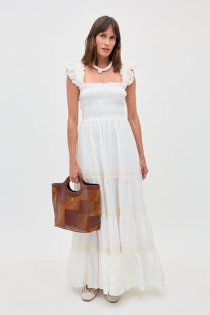 Woman in a long, 70s-inspired ivory nightdress called “Aubrie” with ruffle details holds a brown leather handbag. She wears a chunky white necklace and white shoes, standing against a plain white background.