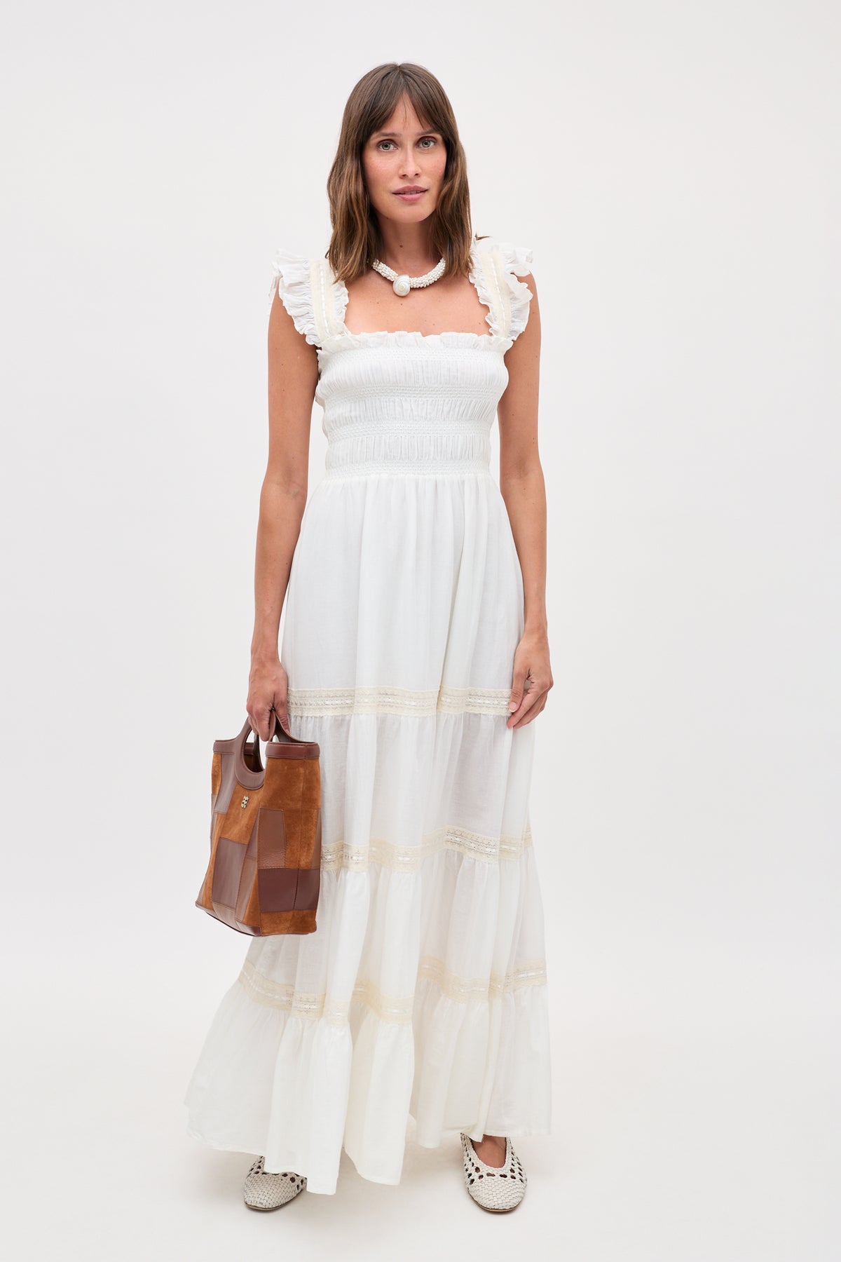 A woman with straight brown hair stands against a plain light background, wearing the Aubrie - Ivory long white ruffled dress inspired by 70s nightwear. She accessorizes with a chunky necklace, white flats, and carries a brown handbag.