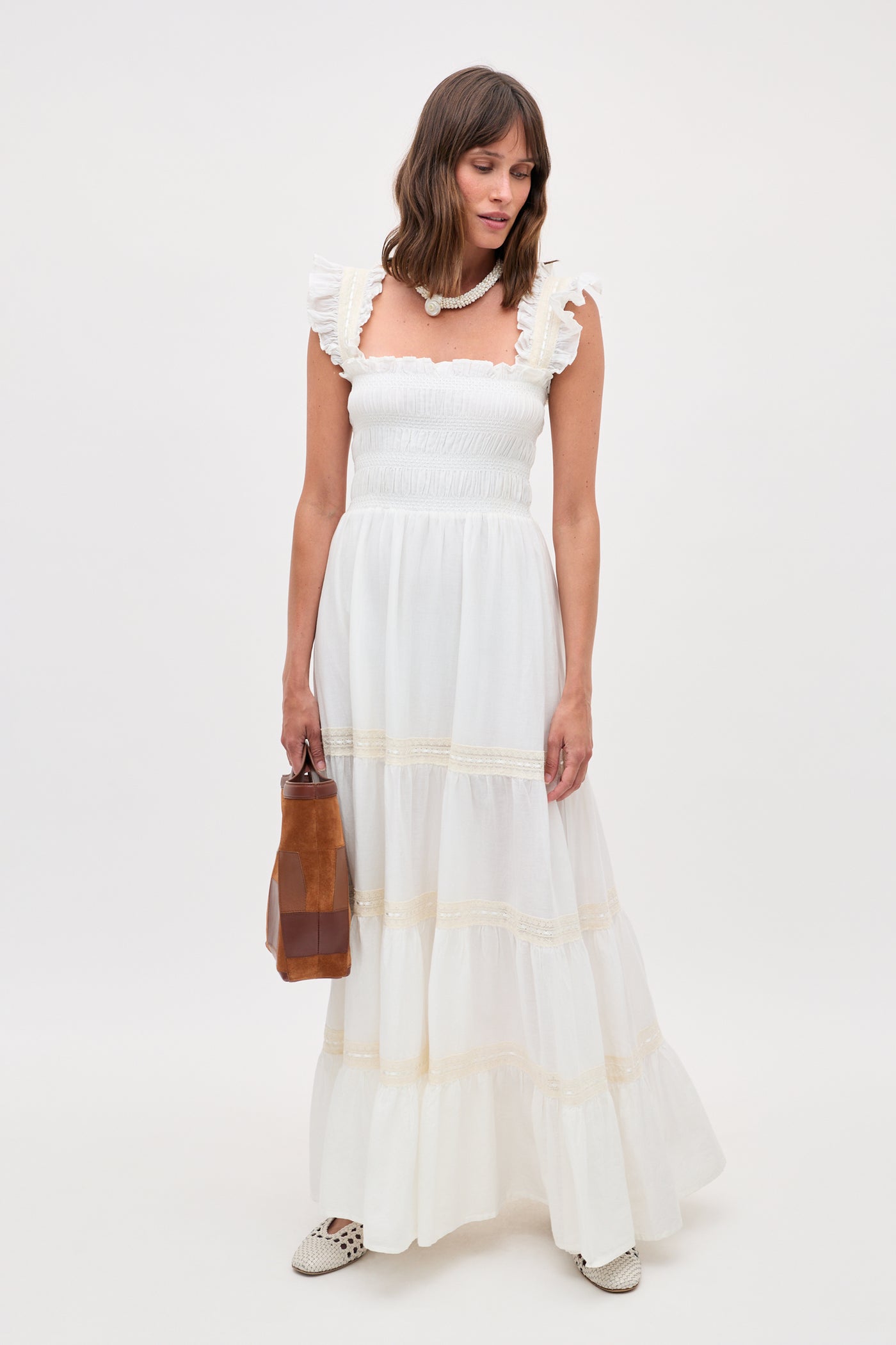 A woman stands against a plain light background wearing the Aubrie - Ivory nightdress, featuring white ruffled details and tiered maxi layers. She holds a brown handbag, accessorizes with a necklace, and completes her look with white cutout shoes.